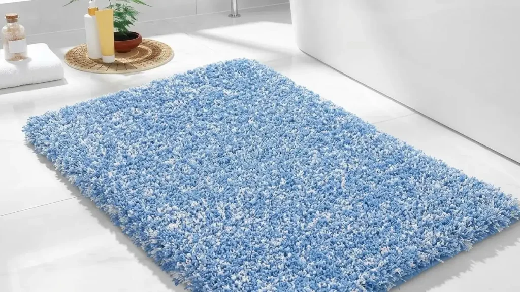 A thick, fluffy blue and white shag bath mat placed next to a modern white bathtub on a bright, sunny marble tile floor with spa accessories nearby.