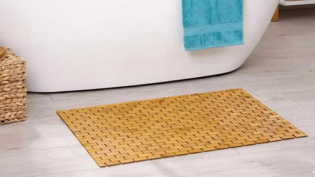 A rectangular slatted bamboo bath mat on a light wood floor in a bathroom, positioned next to a white freestanding bathtub with a turquoise towel. A woven basket sits to the side.