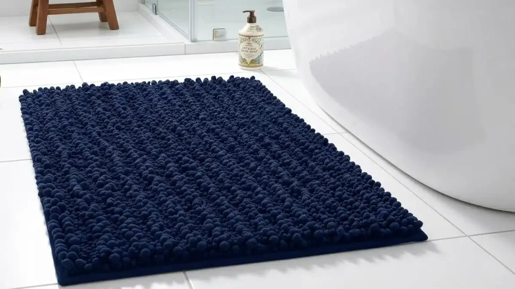 A fluffy, deep navy blue chenille bath mat placed on a white tiled floor next to a modern freestanding bathtub in a clean bathroom.