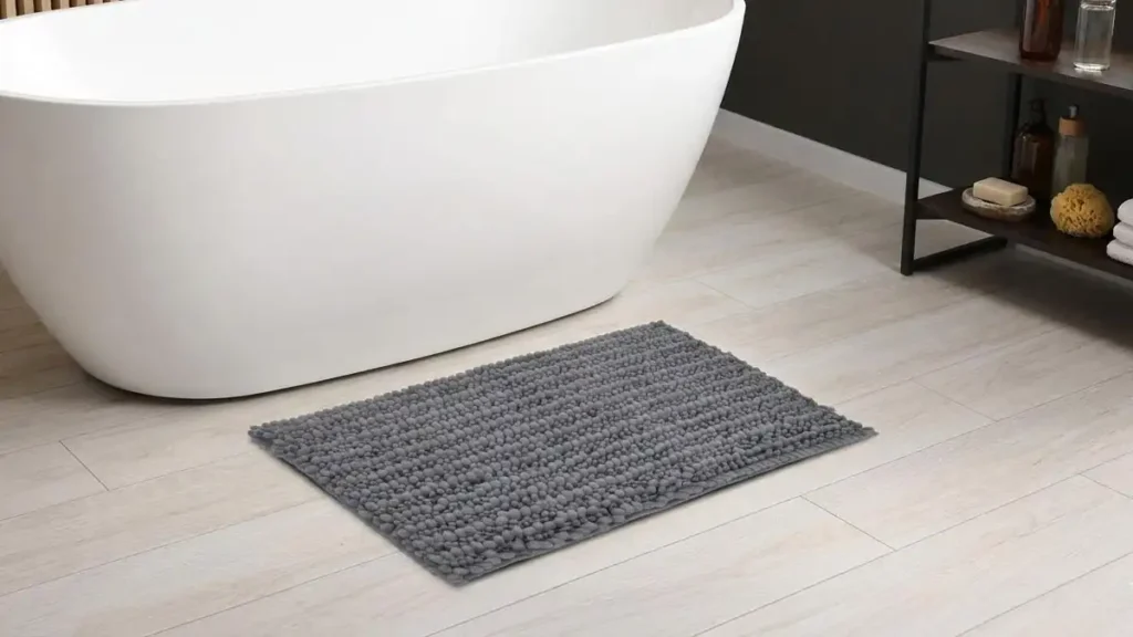 Plush gray polyester microfiber bath mat on a light wood floor next to a modern freestanding white bathtub in a sunlit bathroom.