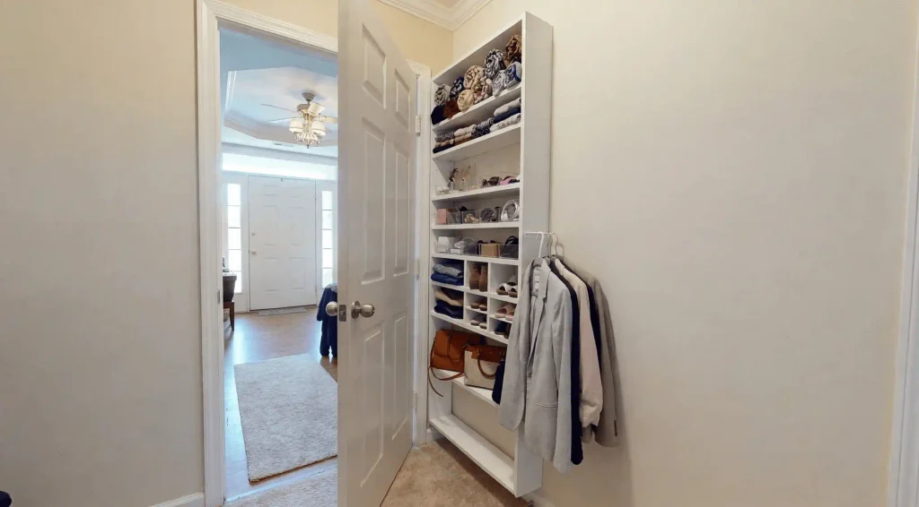 A slim white over-the-door shelf unit mounted behind a bedroom door holding folded scarves, shoes, accessories, handbags, and hanging jackets.